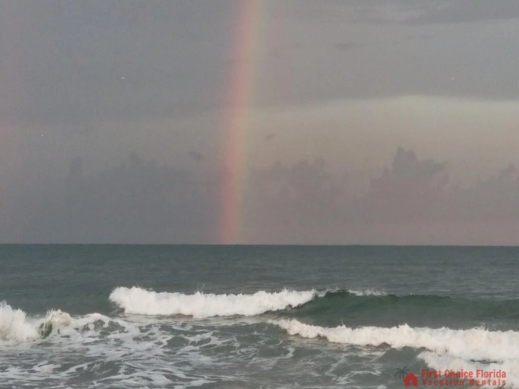 View of ocean waves rolling toward shore under a cloudy sky with a faint vertical rainbow on the horizon.