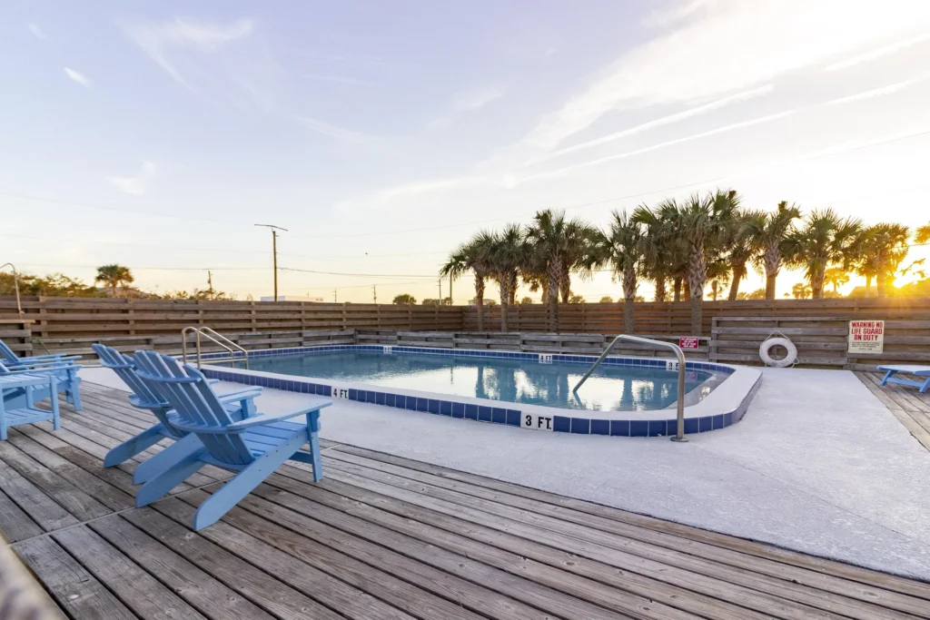 Communal pool area with blue Adirondack chairs on a weathered wood deck, enclosed by a wooden fence and palm trees.