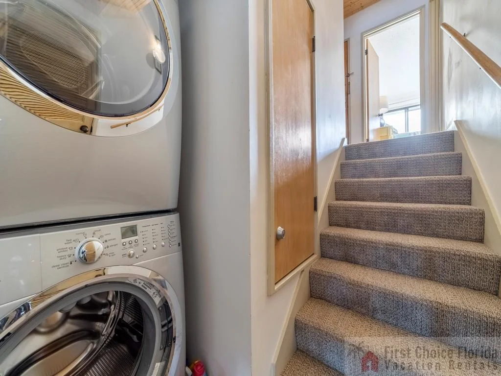 Stacked washer and dryer sit beside carpeted stairs leading to a sunlit upper hallway with wood and white walls.