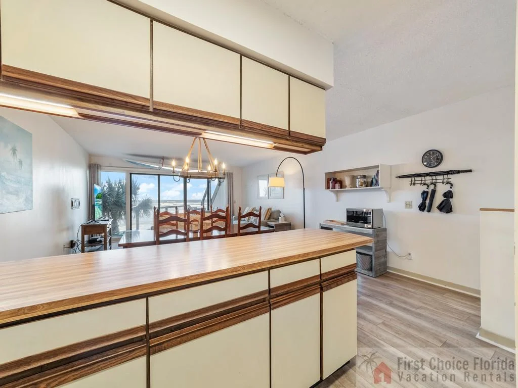 Wood-trimmed kitchen cabinets and countertop frame the view toward a dining area and ocean-facing sliding doors.