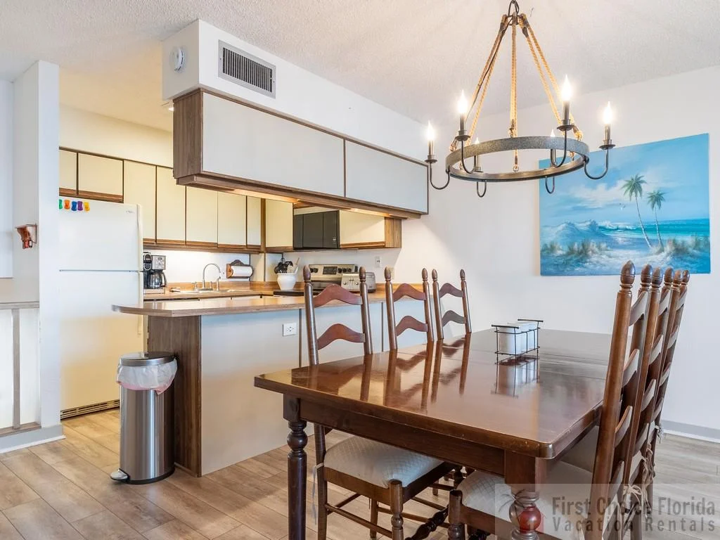 Wood dining table with six ladder-back chairs beneath a wrought-iron chandelier and beach-themed wall art.