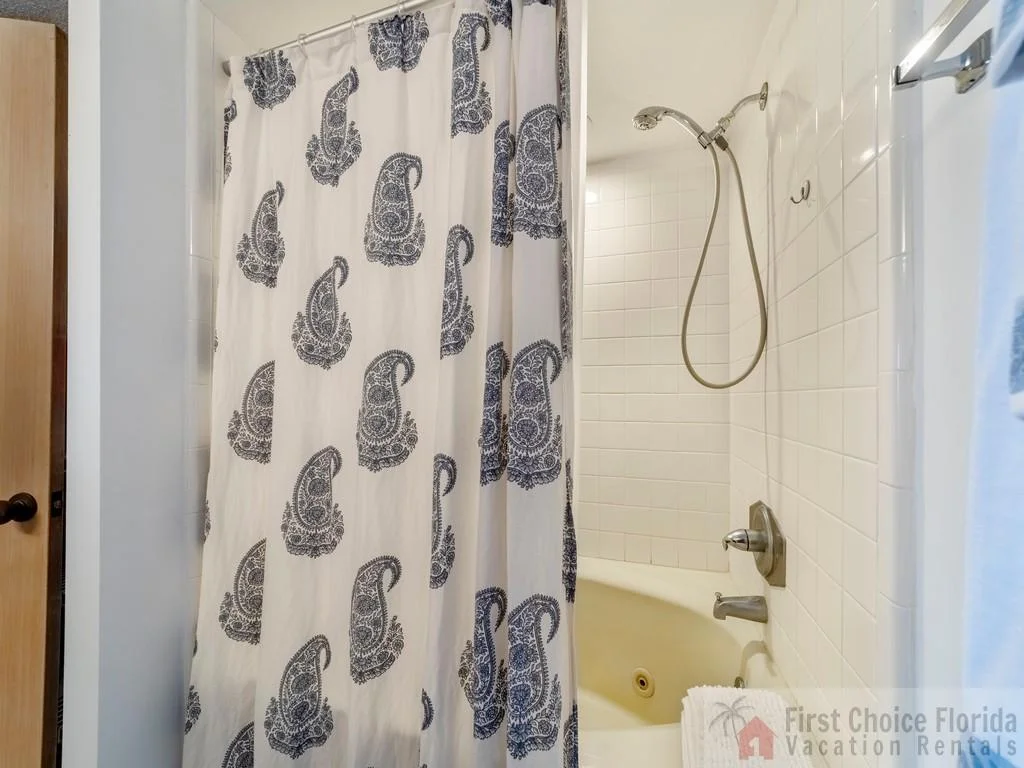 Bathroom shower with white square tiles, chrome fixtures, and a patterned fabric shower curtain.
