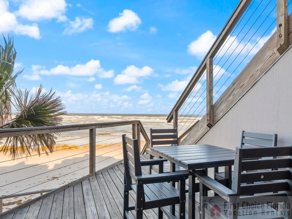 Balcony with dark wood dining set overlooks sandy beach and ocean under a bright blue sky.