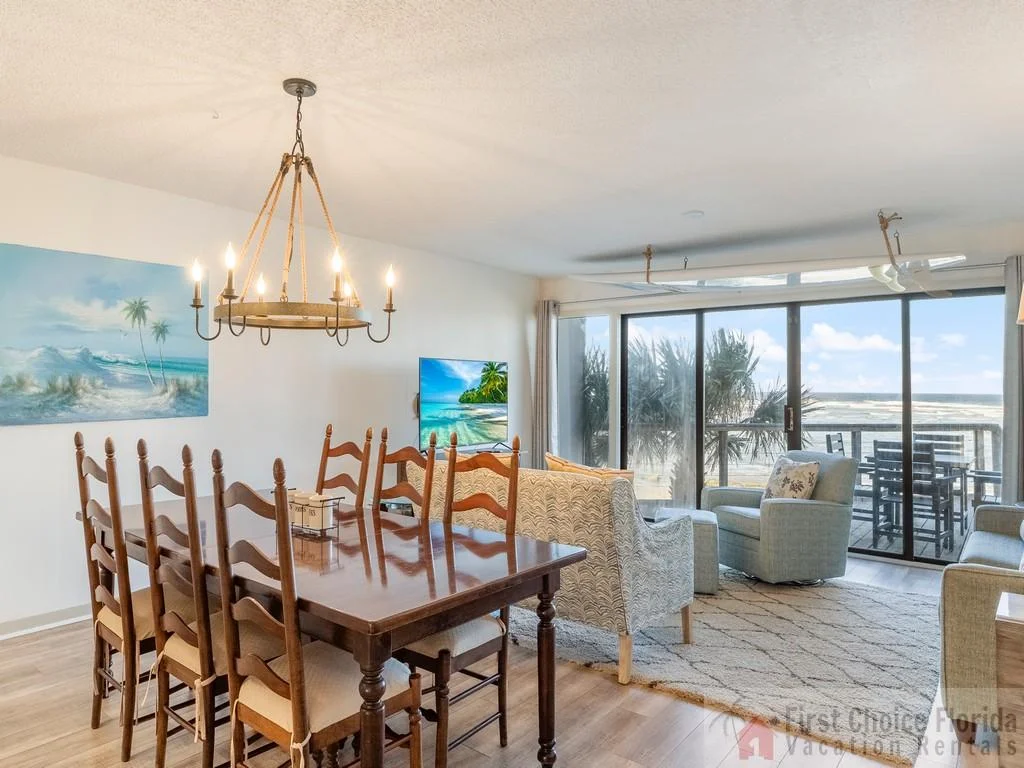 Dining area with a large wooden table and ladder-back chairs, adjacent to a seating area facing sliding glass doors with ocean views.