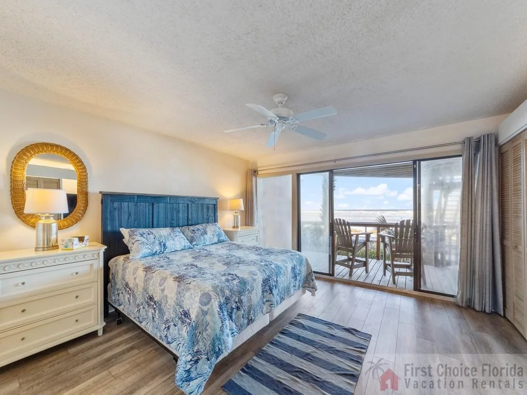 Master bedroom features a king bed with blue patterned bedding, wood floors, and sliding doors to an oceanfront balcony.