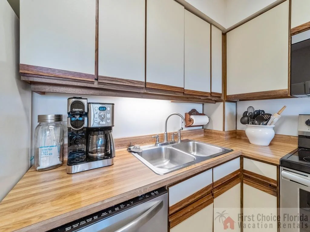 Wood-trimmed cabinets surround a double stainless steel sink, coffee maker, and utensil crock on laminate counters.