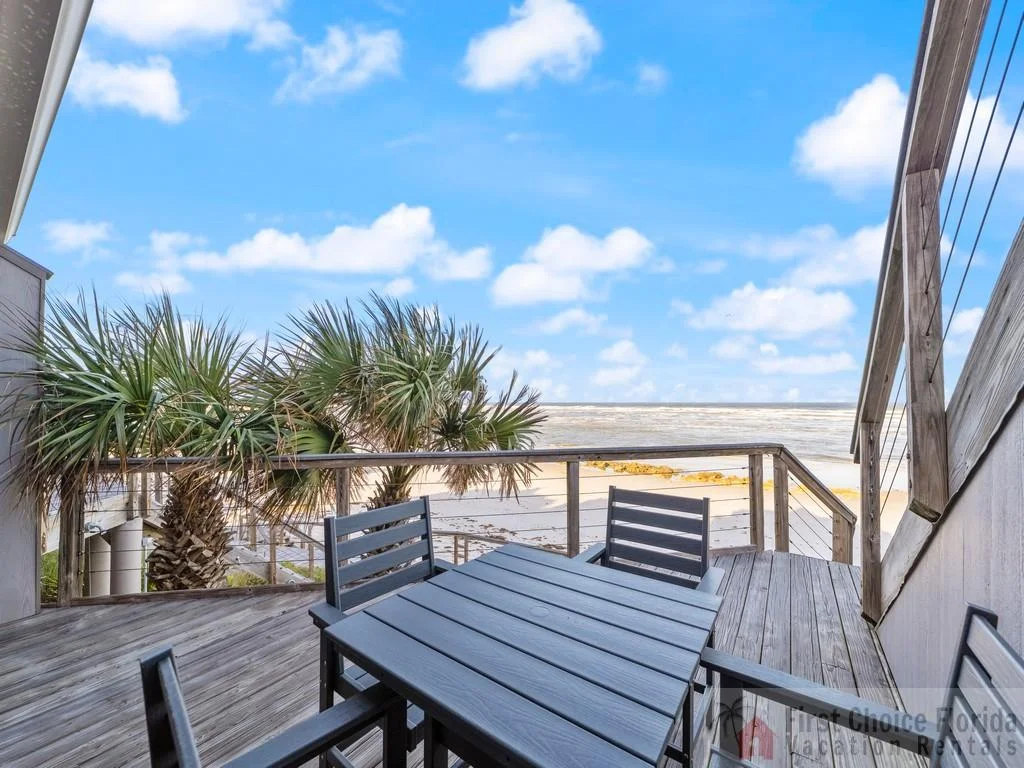 Oceanfront deck with weathered wood planks, a dark gray slatted dining table, and coastal palm trees framing the sandy beach.