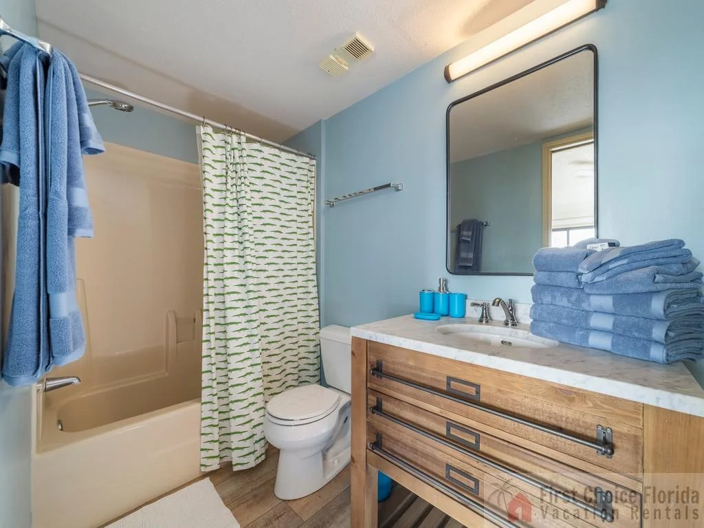 Bathroom with light blue walls, wood vanity topped with marble, and a tub-shower combination with patterned curtain.