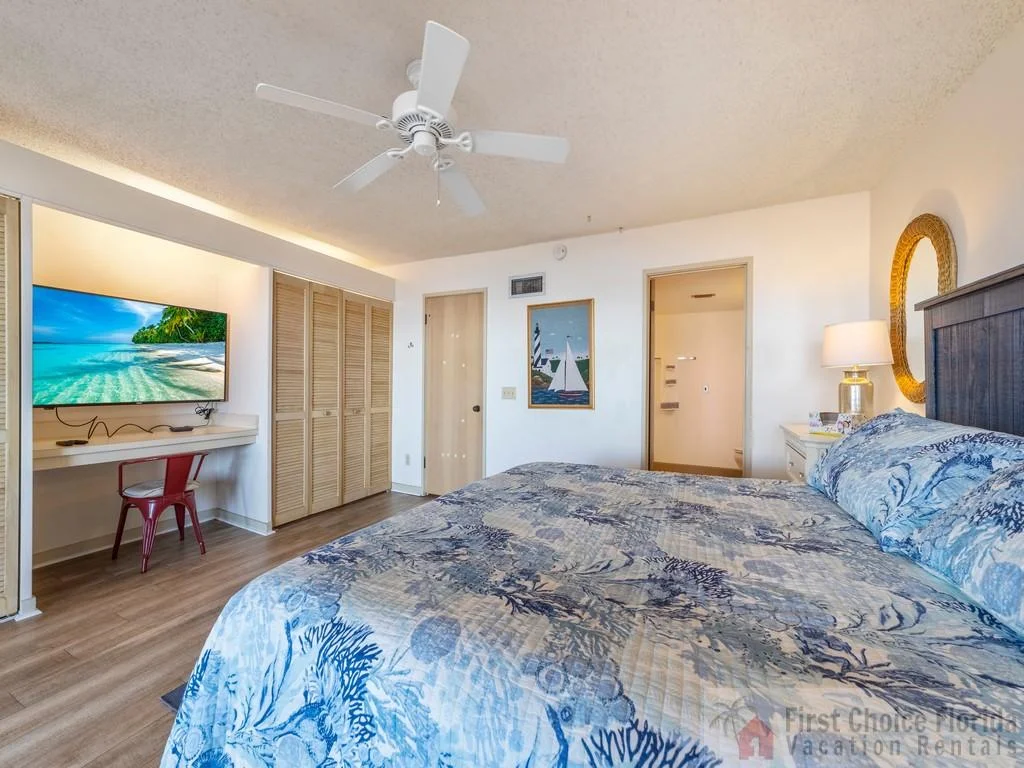 Master bedroom with patterned blue bedding, ceiling fan, wall-mounted TV, and built-in desk with red chair.