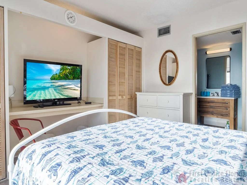 White metal bedframe with a blue seashell quilt faces a wall-mounted TV and a dresser beneath an oval mirror.