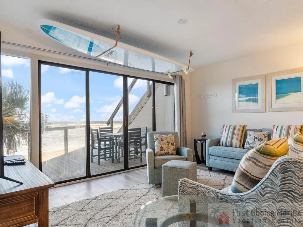 Gray armchair and ottoman face a wooden media console beneath a surfboard mounted above sliding glass doors.