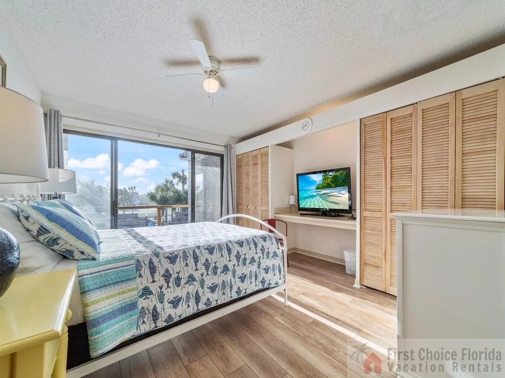 Bedroom with white metal bed frame, coastal-patterned bedding, wood-look flooring, and sliding glass doors to balcony.