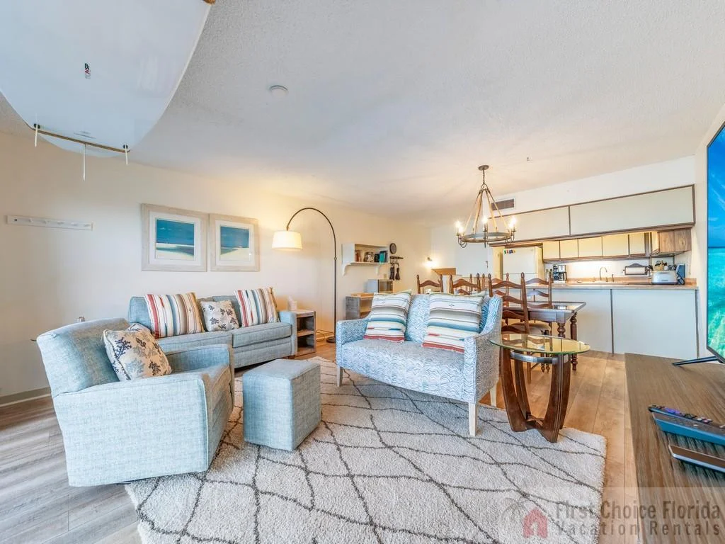 Living area with light gray upholstered seating, patterned rug, wood dining set, and open kitchen in the background.