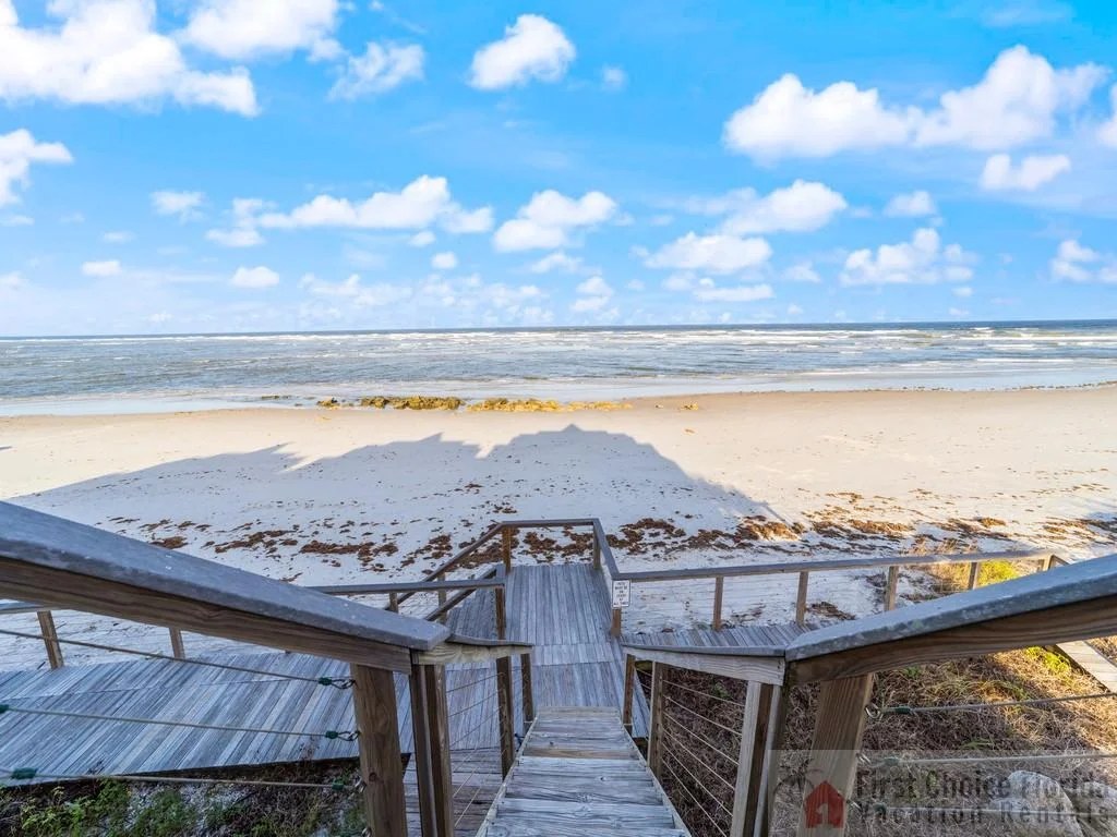 Wooden stairs with cable railings lead down to a sandy beach under a partly cloudy blue sky.