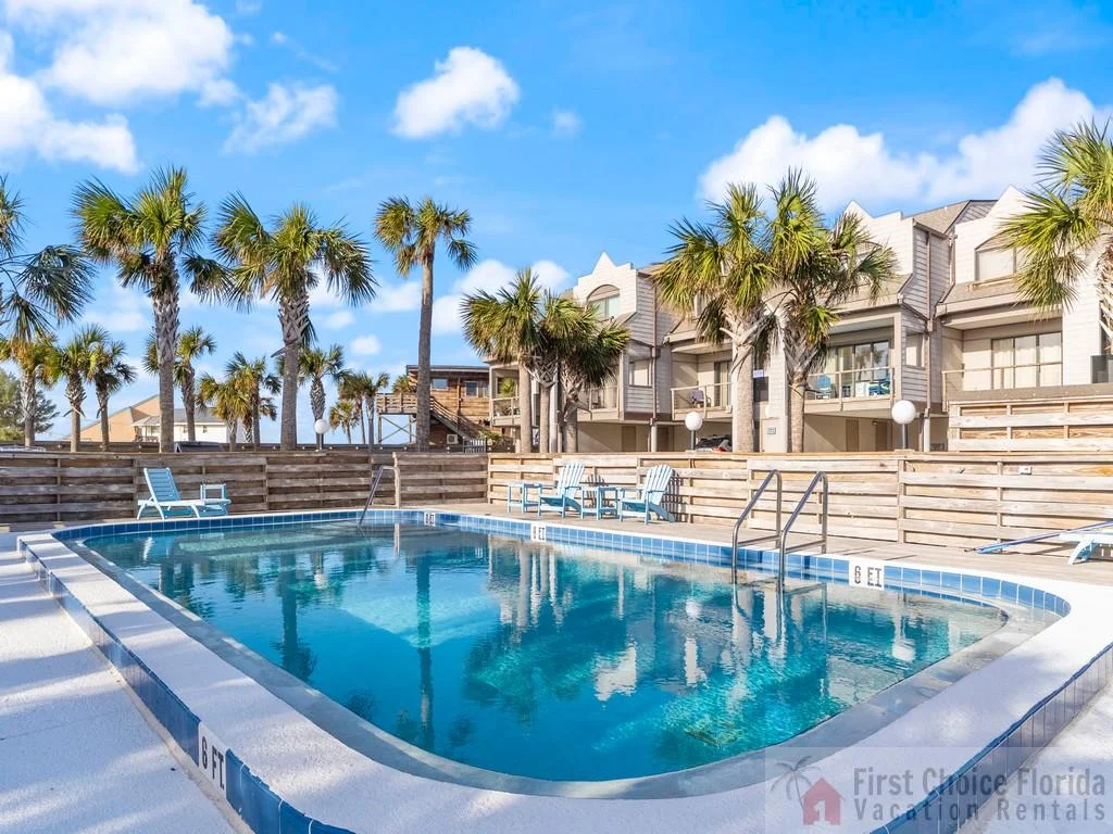Outdoor pool area with blue tile edging, white concrete deck, and Adirondack chairs under palm trees.