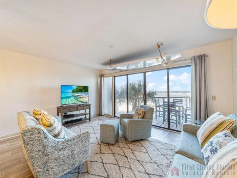 Sliding glass doors open to a balcony with dark wood dining furniture and ocean views beyond the living room seating.