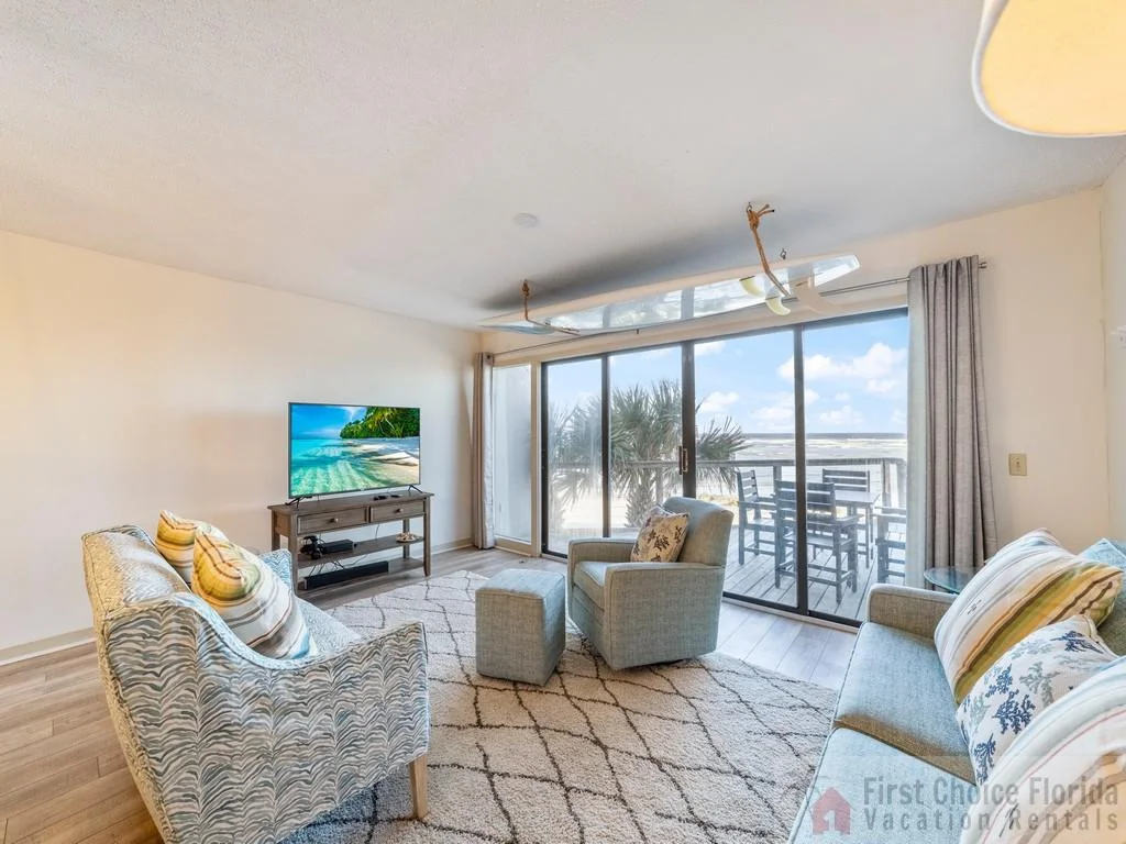 Sliding glass doors open to a balcony with dark wood dining furniture and ocean views beyond the living room seating.