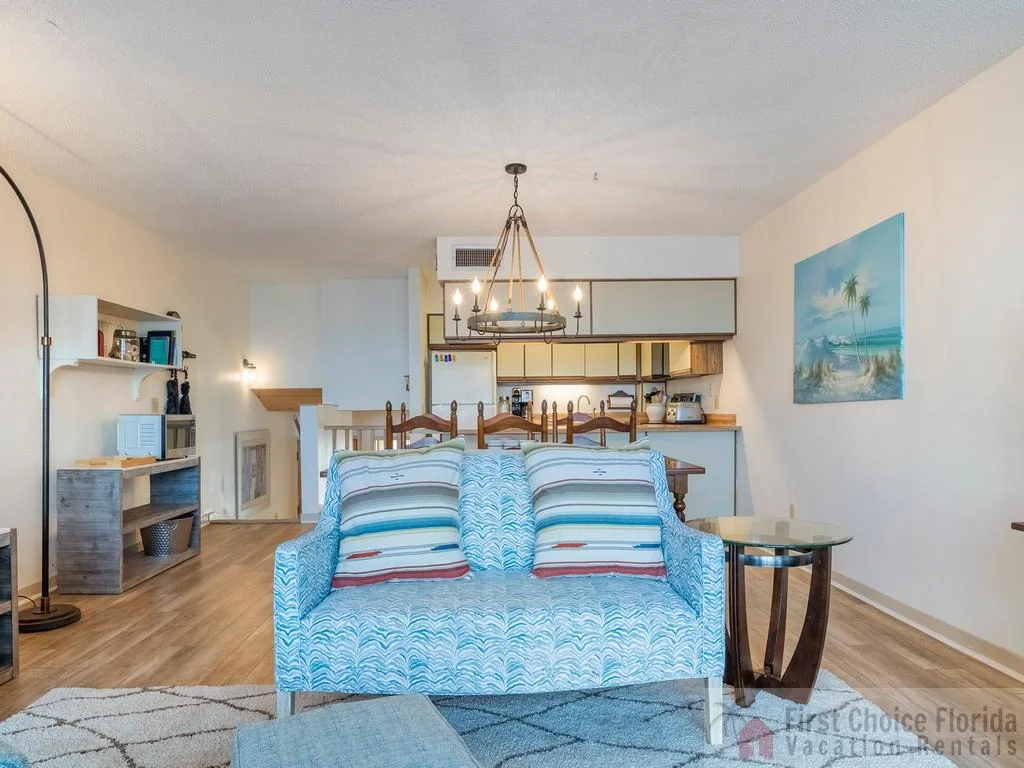 Light wood flooring extends through the living and dining areas, centered by a patterned loveseat and round glass side table.