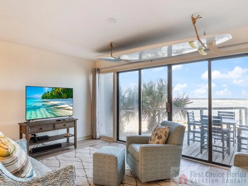 A surfboard hangs from the ceiling above a gray armchair and ottoman by sliding glass doors leading to a balcony with ocean views.