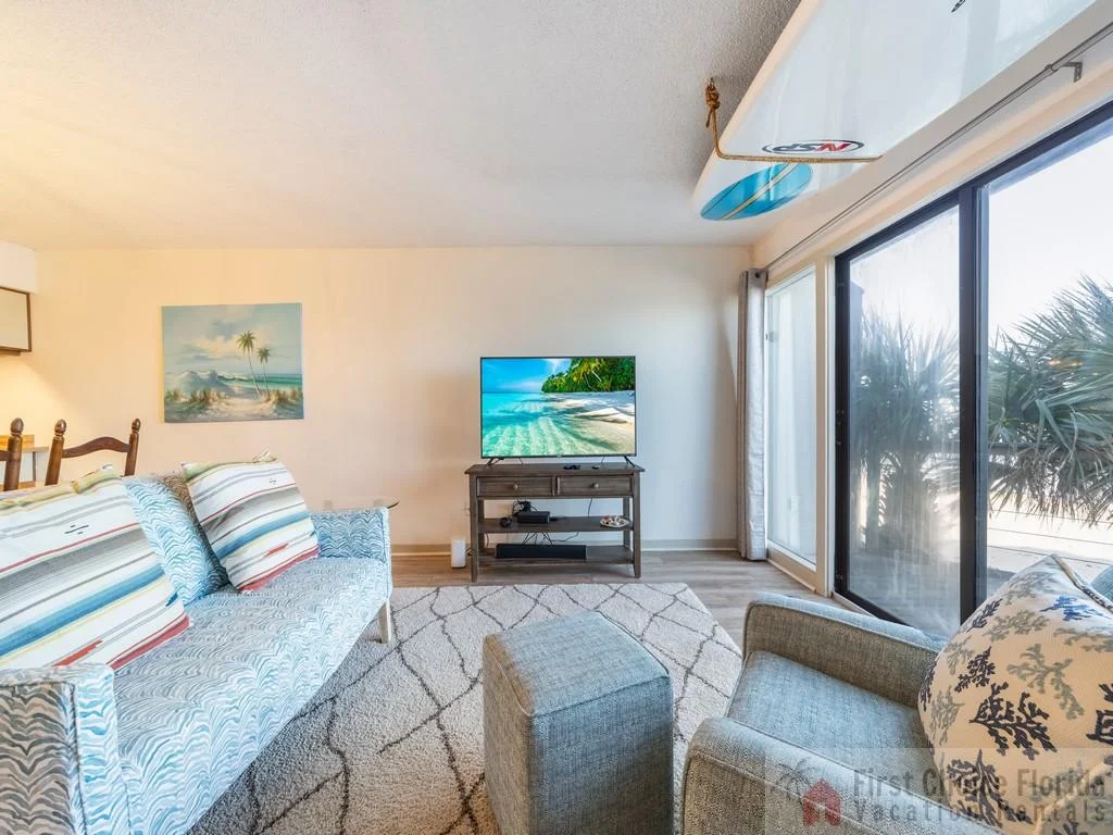 Light wood floors anchor the seating area with patterned sofa, gray armchair, and a surfboard mounted near sliding glass doors.