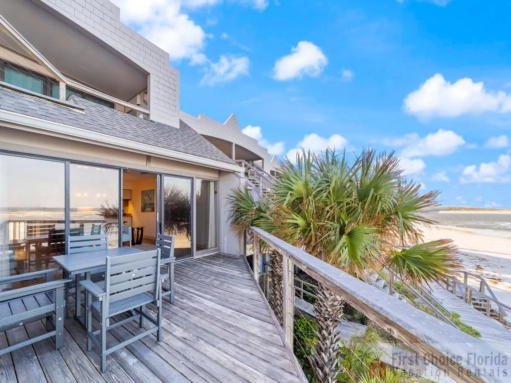 Weathered wood balcony with gray dining set overlooks palm trees, sandy beach, and ocean under blue sky.