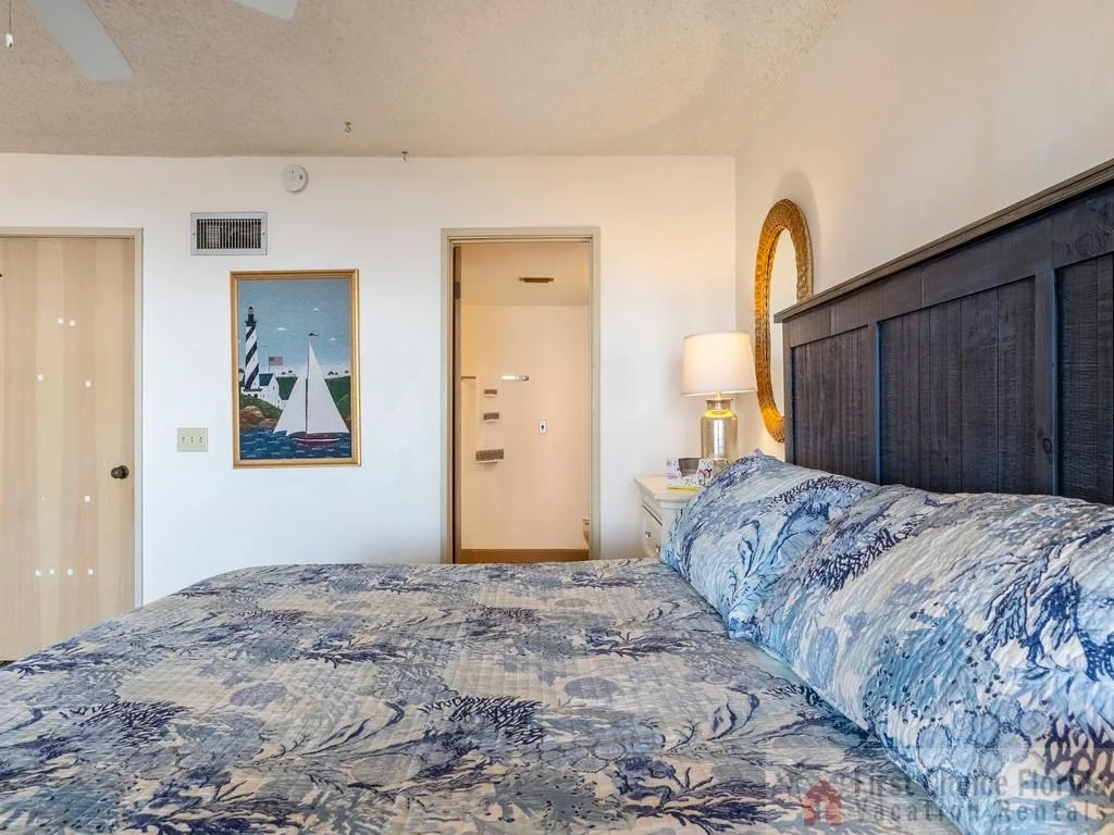 Master bedroom with dark wood headboard, seashell-patterned bedding, woven mirror, and lamp on white nightstand.