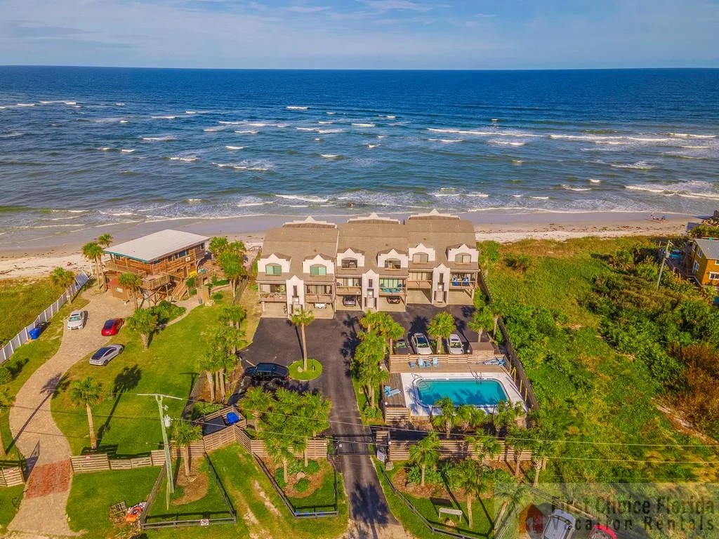 Oceanfront condo complex with rooftop balconies, parking area, fenced pool, and sandy beach access.