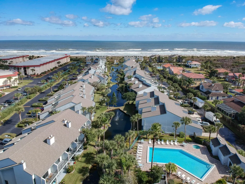 Aerial view of condo complex with lagoon, palm trees, pool, and the ocean beyond under a partly cloudy sky.