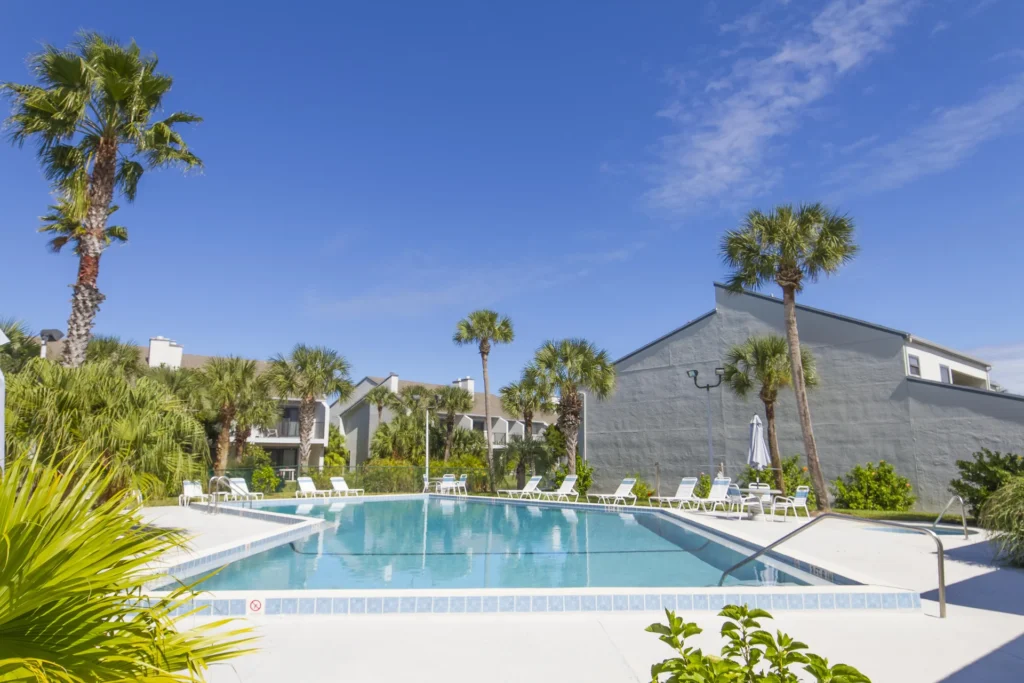 Outdoor swimming pool surrounded by white lounge chairs and tropical palm trees under a clear blue sky.