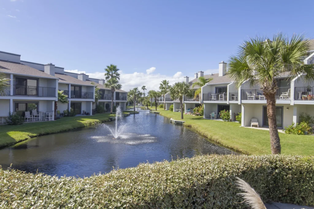 Lagoon view with water fountain framed by two-story condos, green lawns, palm trees, and clear blue sky.