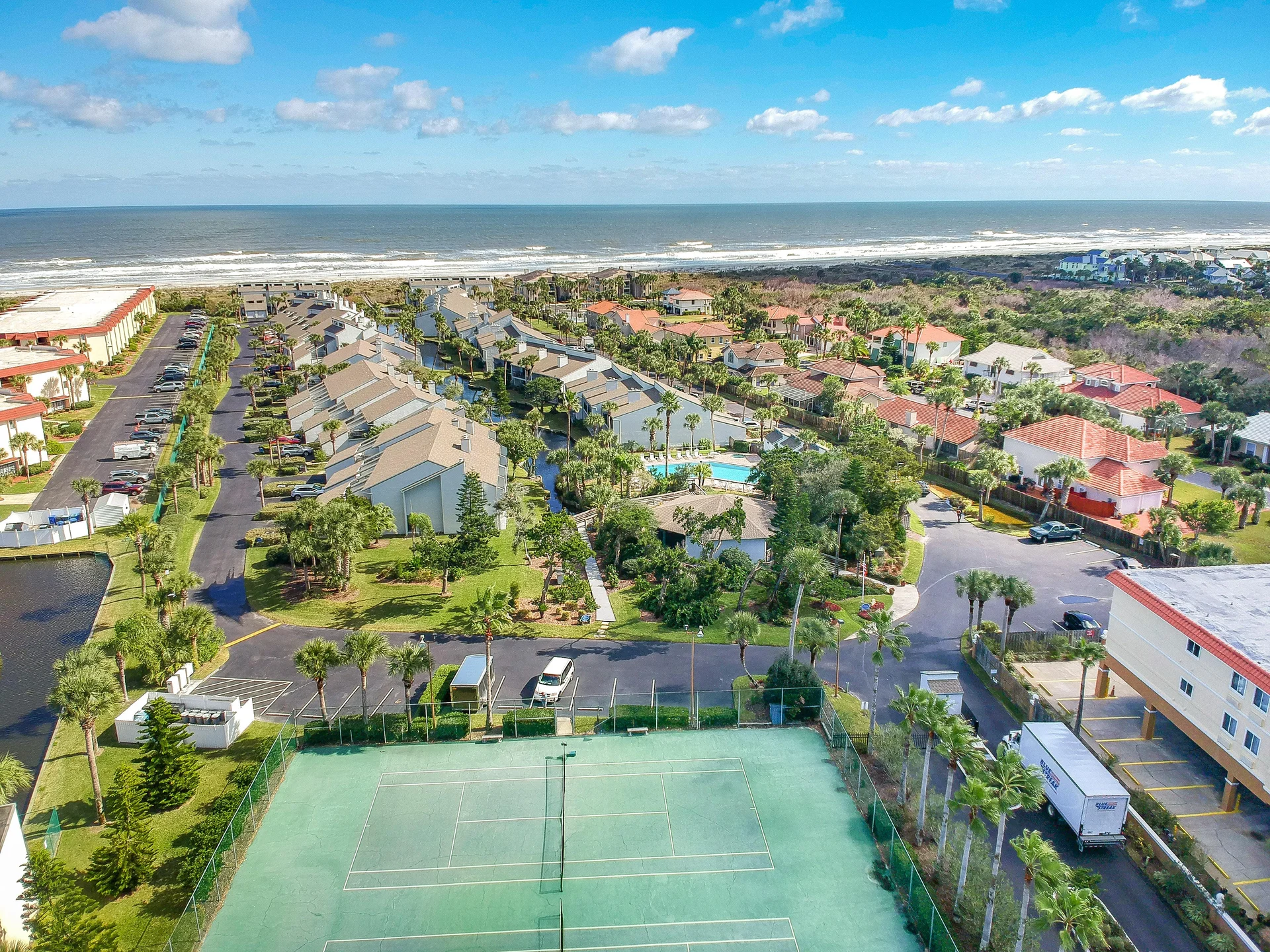 Tennis courts with green surfaces and netting sit at the edge of the condo complex near palm trees and ocean views.