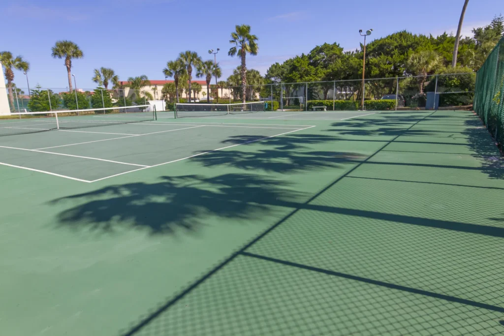 Green tennis court with white boundary lines, surrounded by chain-link fencing and palm trees under a clear sky.