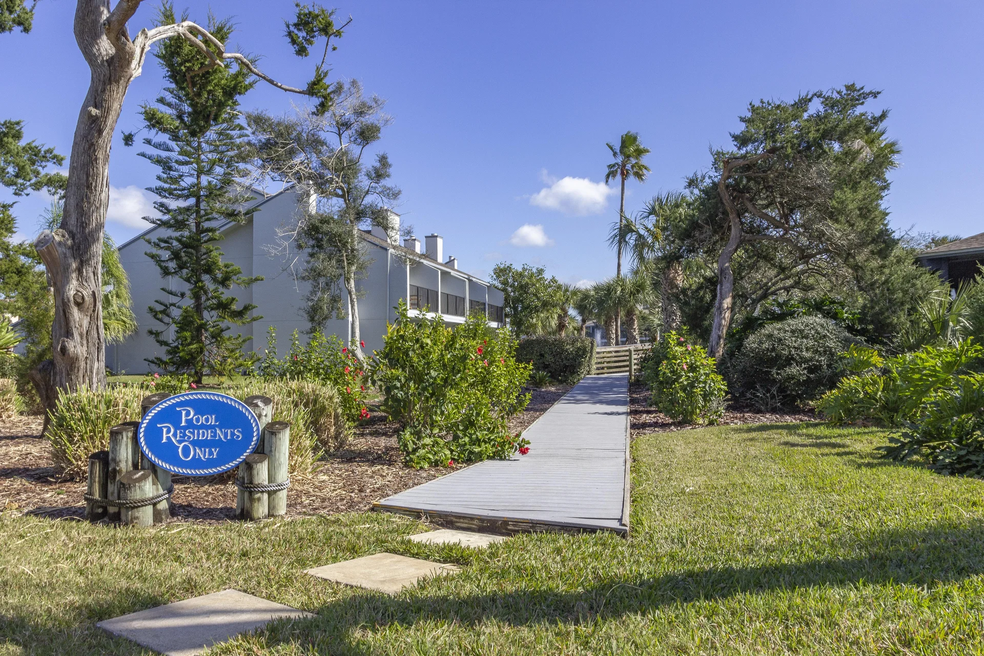 Wooden boardwalk path leads through landscaped greenery and blooming hibiscus toward condo buildings under clear blue sky.