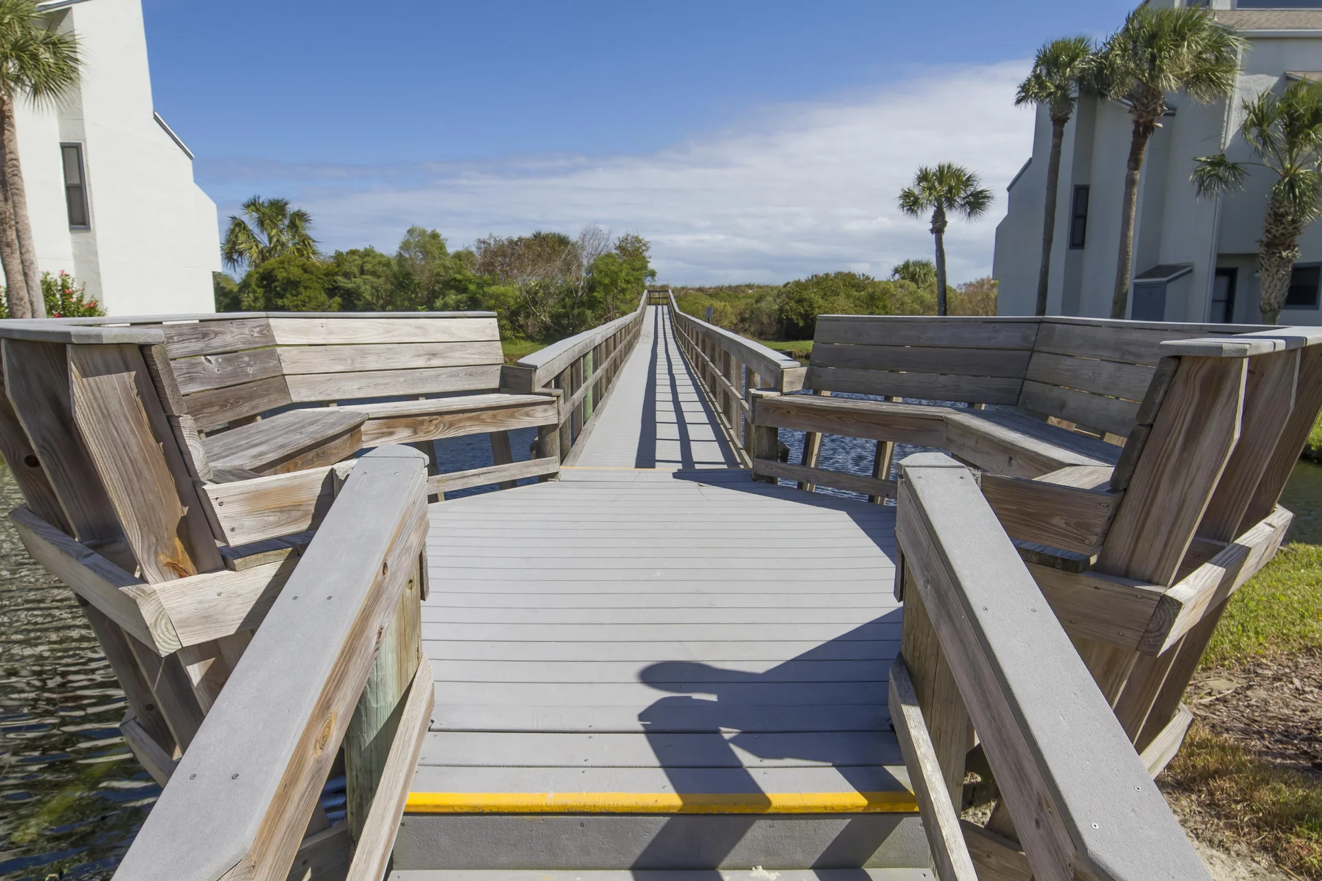 Wooden seating benches flank a gray boardwalk leading over a lagoon, surrounded by palm trees and greenery.