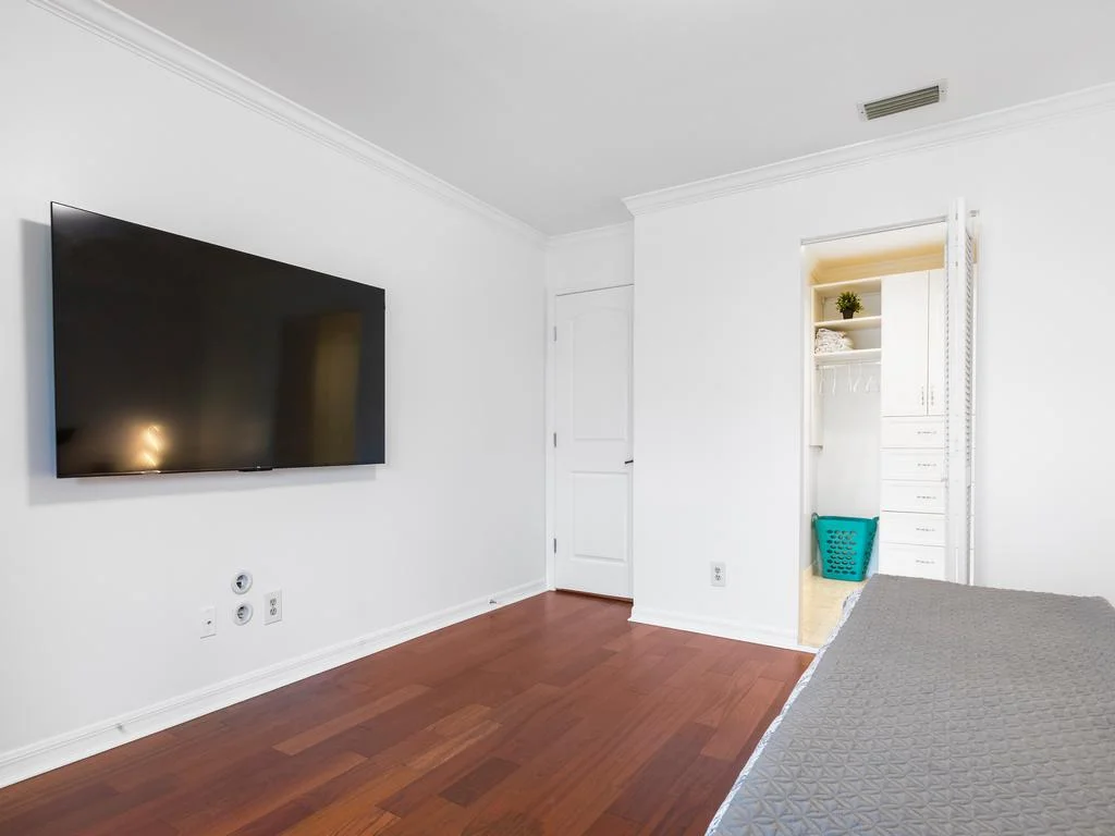 Wall-mounted flat-screen TV faces a gray quilted bedspread on polished hardwood floors in the bedroom.