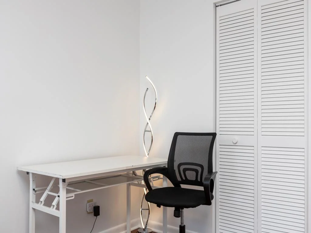 Work space with a white metal desk, black mesh office chair, and sculptural LED floor lamp by louvered closet doors.