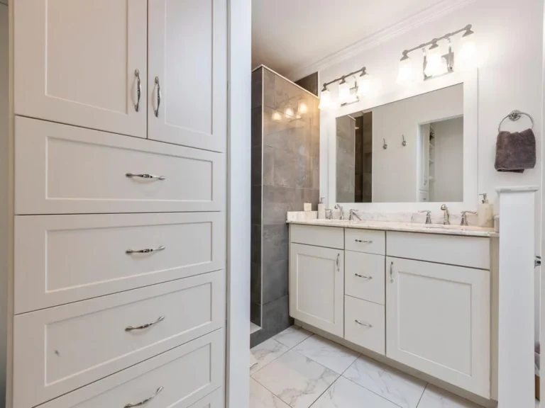White cabinetry and marble countertop frame the double-sink vanity beside a glass-enclosed shower with gray tile.