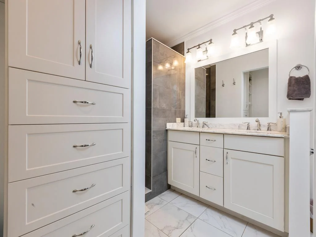 White cabinetry and marble countertop frame the double-sink vanity beside a glass-enclosed shower with gray tile.