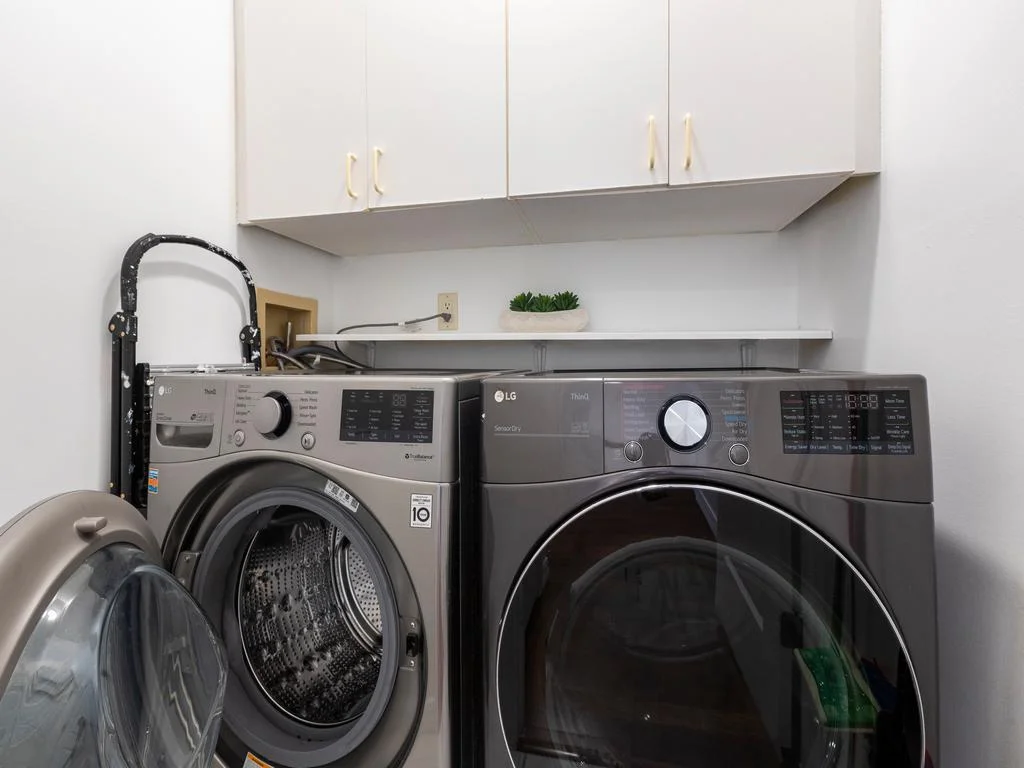 Front-loading washer and dryer in a compact laundry nook beneath white storage cabinets and a small shelf with decor.