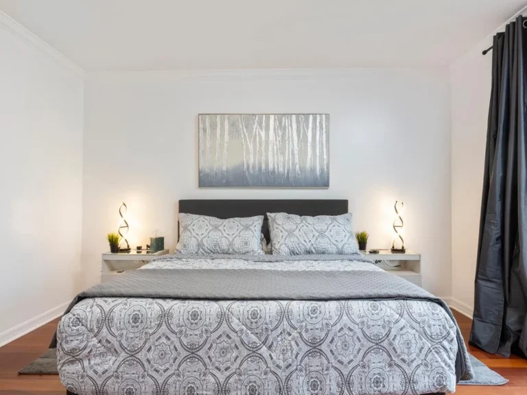 King bed with patterned bedding between white nightstands and modern twisted lamps, dark curtains to the side.