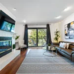 Living room features a dark leather sectional, glass coffee table, and a wall-mounted TV above a white tiled fireplace.