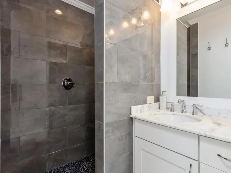 Walk-in shower with large gray tiles and pebble stone floor beside a white vanity topped with marble countertop.