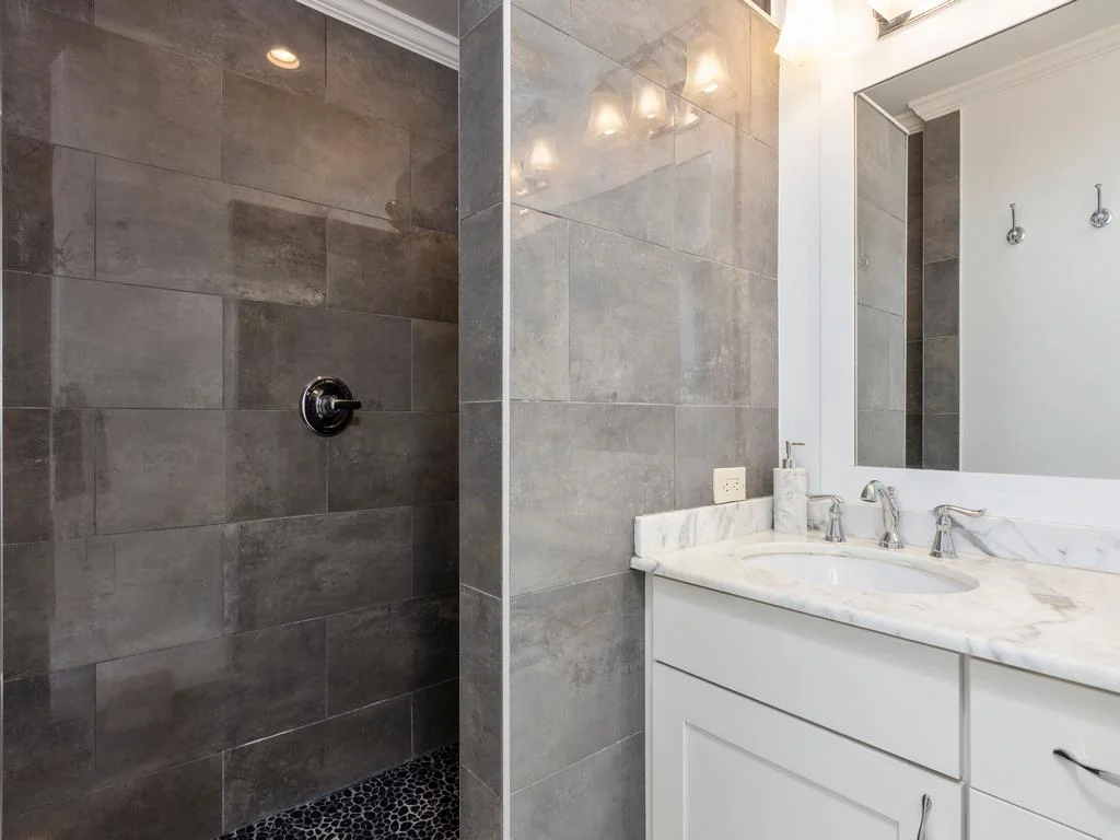 Walk-in shower with large gray tiles and pebble stone floor beside a white vanity topped with marble countertop.