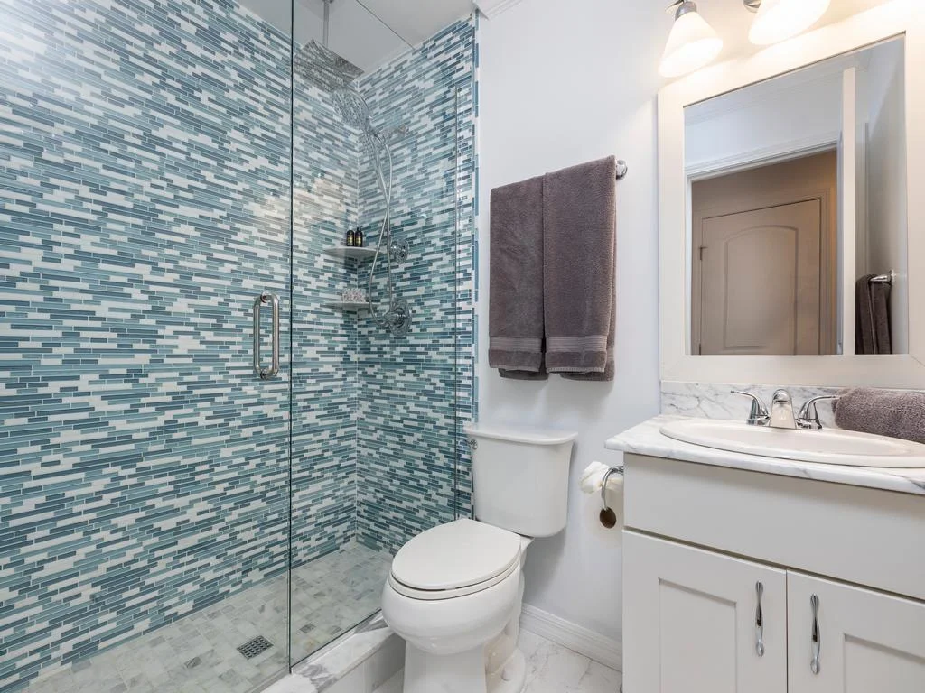Glass-enclosed shower features blue and white mosaic tiles, with white vanity and marble countertop nearby.