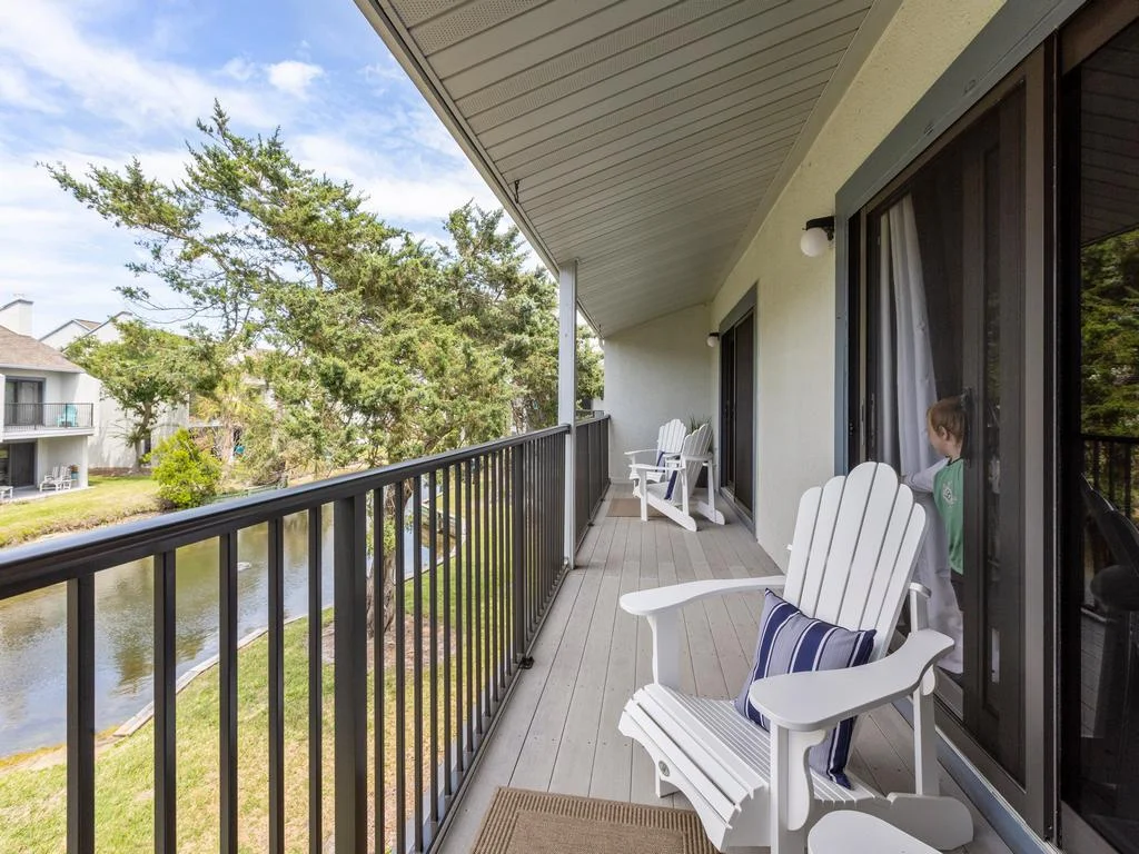 Covered balcony with white Adirondack chairs and striped pillows overlooking a narrow lagoon and trees.