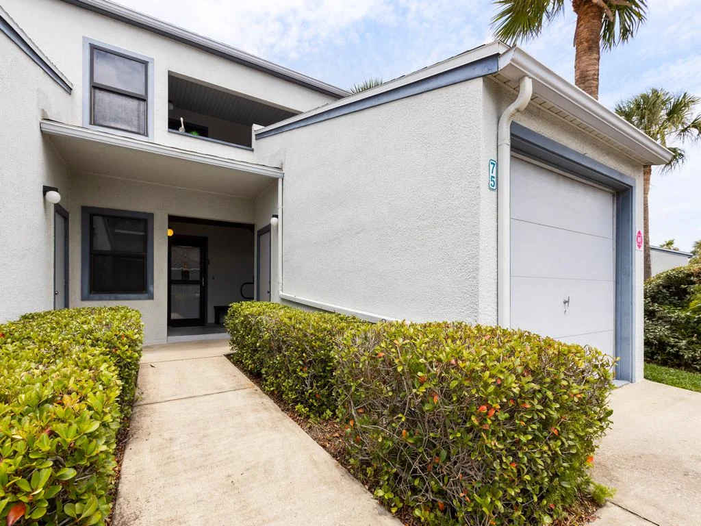 Concrete walkway bordered by trimmed green hedges leads to the white stucco condo entrance.