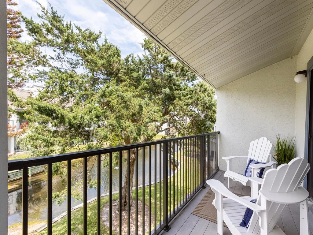 Covered balcony with white Adirondack chairs overlooks a tree-lined lagoon and grassy bank.