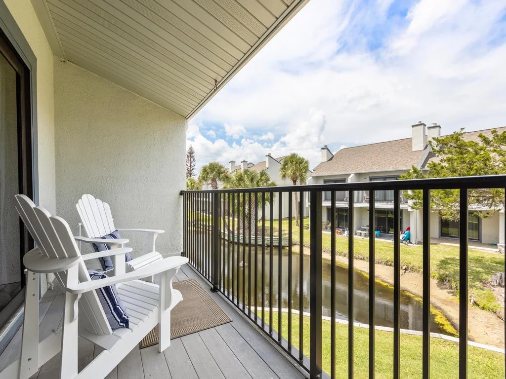 Two white Adirondack chairs with striped cushions sit on a covered balcony overlooking a narrow lagoon and neighboring condos.