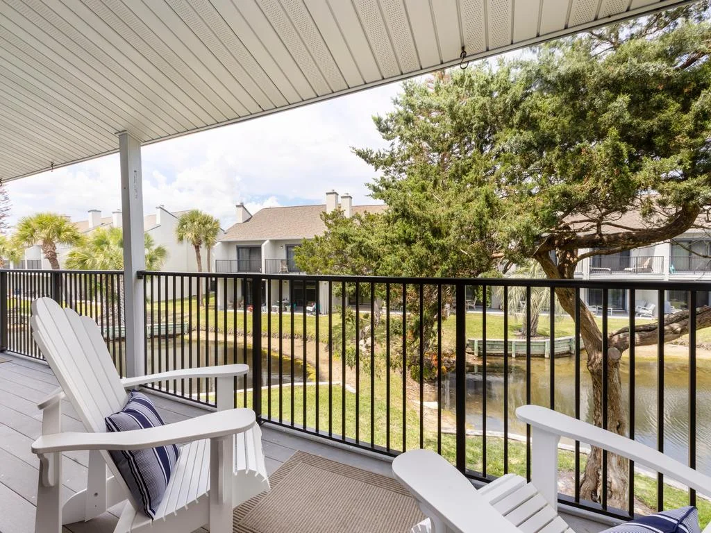 White Adirondack chairs with striped pillows face a black metal railing overlooking a tranquil lagoon and trees.