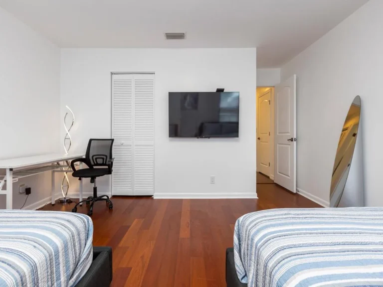 Wall-mounted TV faces two twin beds with blue-striped bedding on polished hardwood floors.